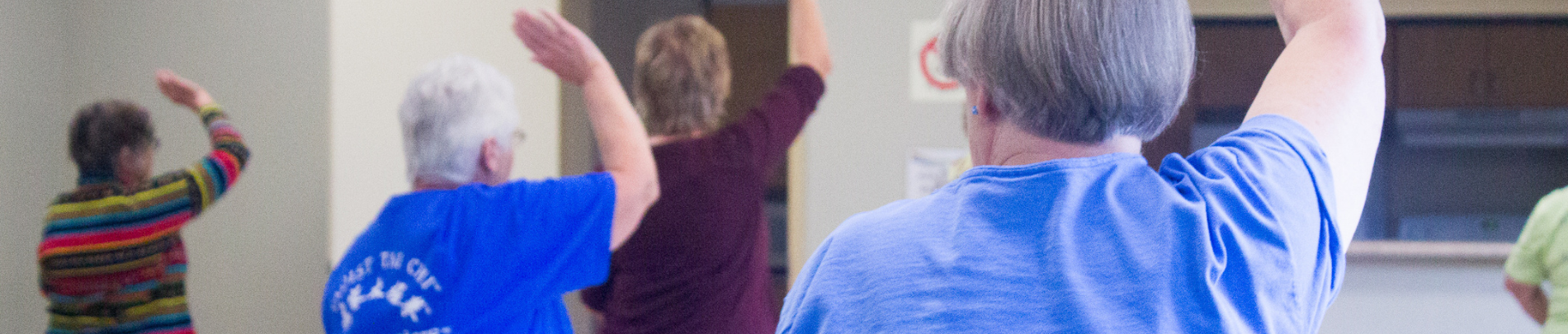A group of older adults participating in a fitness class indoors, raising their arms in unison during a low-impact movement session.