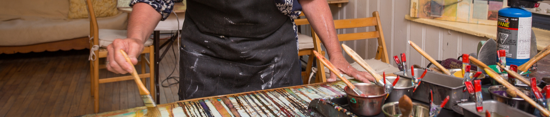 An artist wearing a black apron works on a vibrant painting using a brush, surrounded by art supplies and tools in a cozy studio.