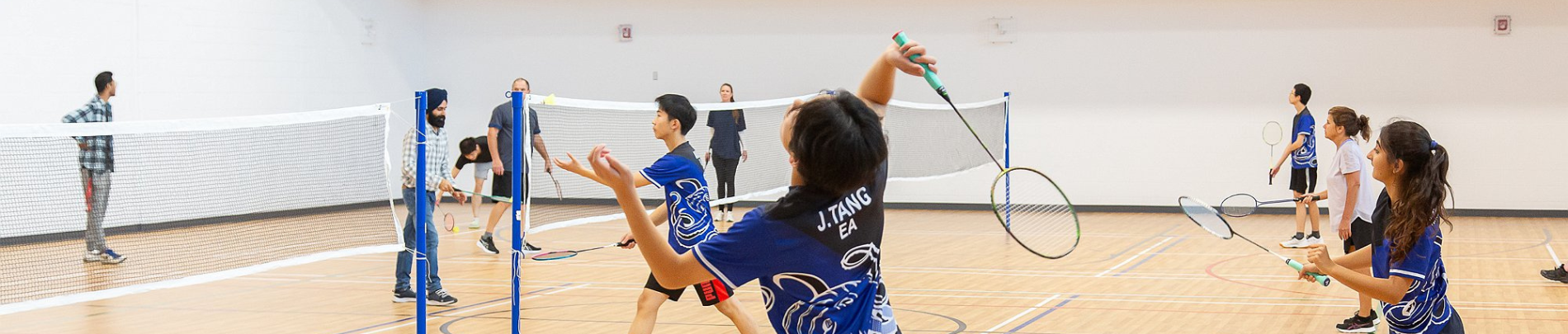 Youth and adults play badminton in a gymnasium, actively engaged in a drop-in match while others watch or wait to play.