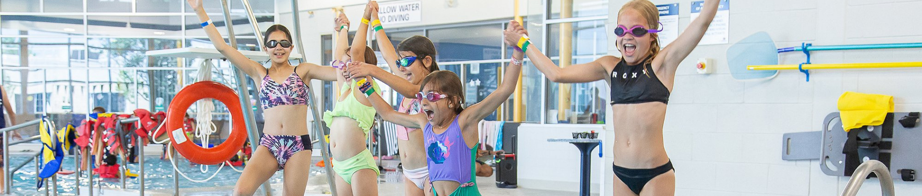 A group of five children wearing swimsuits and swim goggles stand beside an indoor swimming pool with their arms raised. Pool safety equipment, including a bright orange life ring and life jackets, is visible in the background. The aquatic facility is brightly lit with large windows and poolside equipment along the walls.