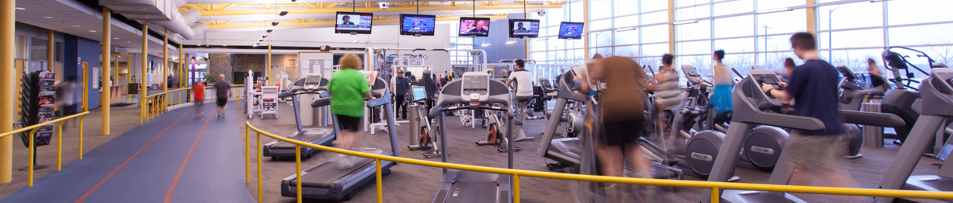 A wide-angle view of a fitness centre with people using treadmills, stationary bikes, and cardio machines along an indoor walking track under natural and artificial lighting.