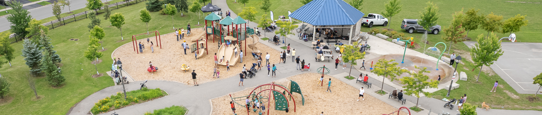 A wide aerial view of a community park with playgrounds, splash pad, pavilion, and residents enjoying various outdoor activities on a sunny day.