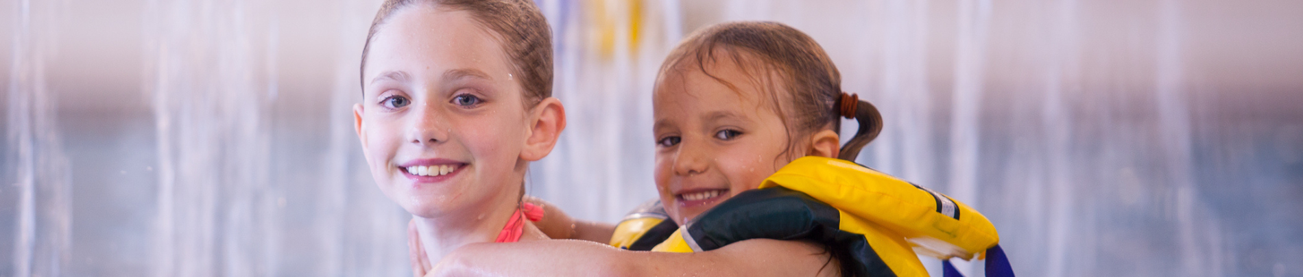 Two smiling children in a swimming pool. One child is wearing a yellow life jacket and hugging the other child from behind, both looking at the camera with water cascading in the background.
