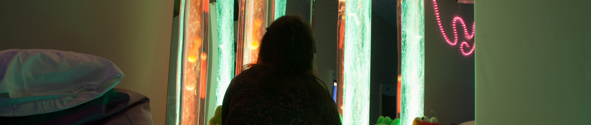A person sits in a dimly lit sensory room, surrounded by glowing bubble tubes and soft ambient lighting designed for relaxation and sensory stimulation.